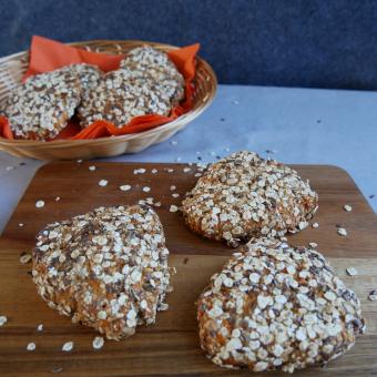 Im Vordergrund liegen auf einem Holzbretchen drei  Brötchen die die Form eines Dreieckes haben und mit Haferflocken bestreut sind. Im Hintergrund sind vier der Karottenecken in eime Brotkorb mit orangener Serviette