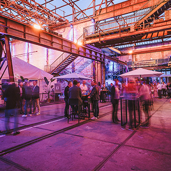 Eine rießige Halle mit vielen Stahlträgern - darin Stehtische mit vielen Menschen.