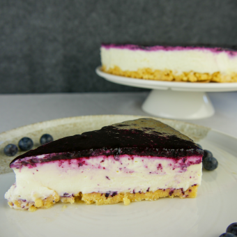 Im vordergrund ist ein Stück Heidelbeer-Cheesecake, ein Cheesecake mit Keksboden und einem Heidelbeerfruchtspiegel. Im Hintergrund steht der ganze Kuchen