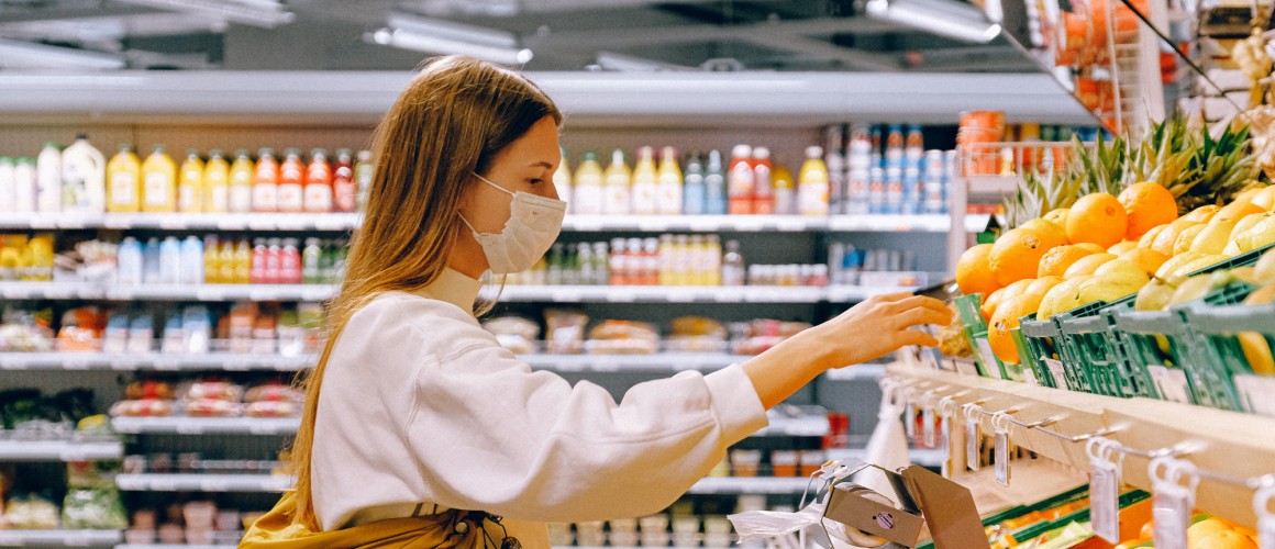 Frau steht mit Mundschutz und Einkaufskorb im Supermarkt vor einem Obstregal
