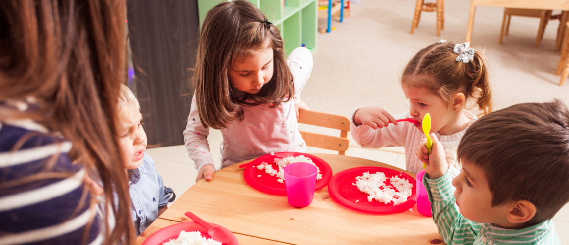 Kitakinder sitzen während des Essens gemeinsam am Tisch. Pädagogische Fachkraft unterstützt die Kinder dabei.