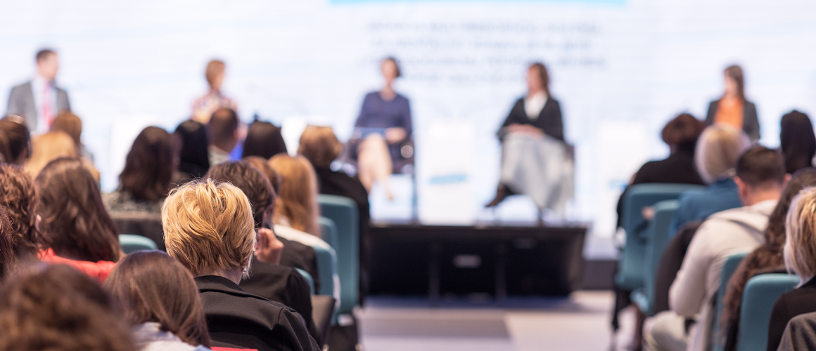 Zuschauer von Hinten vor Bühne auf der 5 Redner sitzen. Dahinter eine Präsentation
