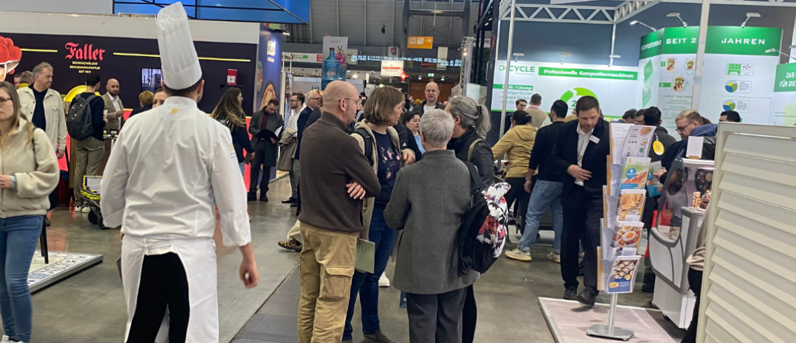 Koch sieht man von hinten mit Kochmütze spazierrend über die Messe