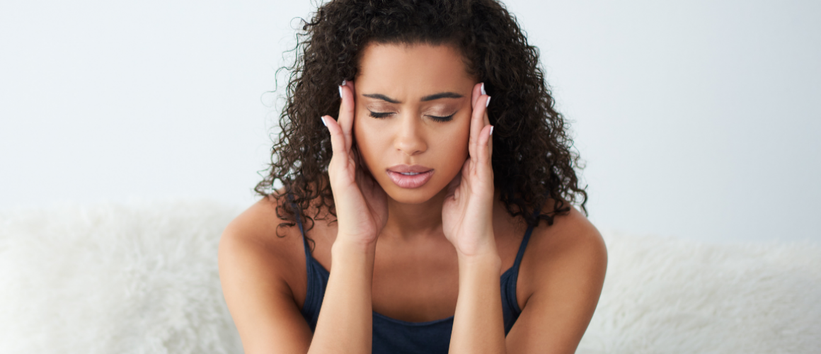 Eine Frau sitzt auf dem Bett, wirkt erschöpft und leidet unter Kopfschmerzen.