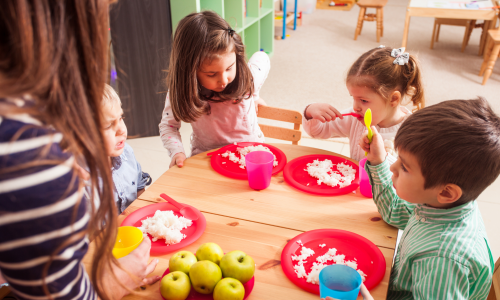 Kitakinder sitzen während des Essens gemeinsam am Tisch. Pädagogische Fachkraft unterstützt die Kinder dabei.