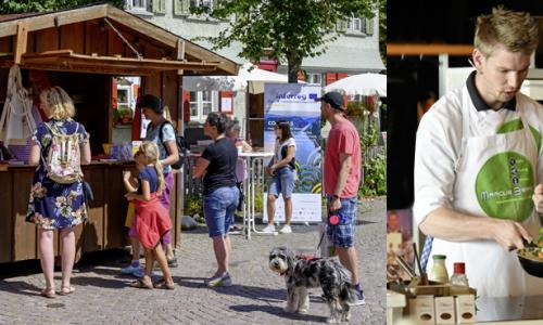 Markstand mit Marktbesuchern, ein Koch der Gemüse in einer Pfanne wendet, Albhornbläser