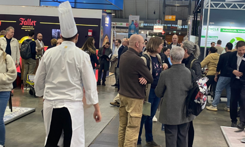 Koch sieht man von hinten mit Kochmütze spazierrend über die Messe