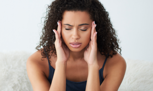 Eine Frau sitzt auf dem Bett, wirkt erschöpft und leidet unter Kopfschmerzen.